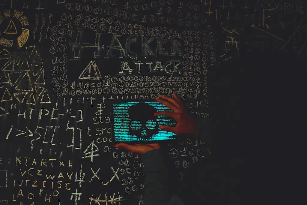 Tafel mit Schlagwörtern wie „Hacker“ und „Attack“ in großer Schrift. Im Vordergrund ein Laptop mit dem Bild eines Hackers auf dem Bildschirm – Symbolbild für Cyberangriff oder Hacking-Attacke